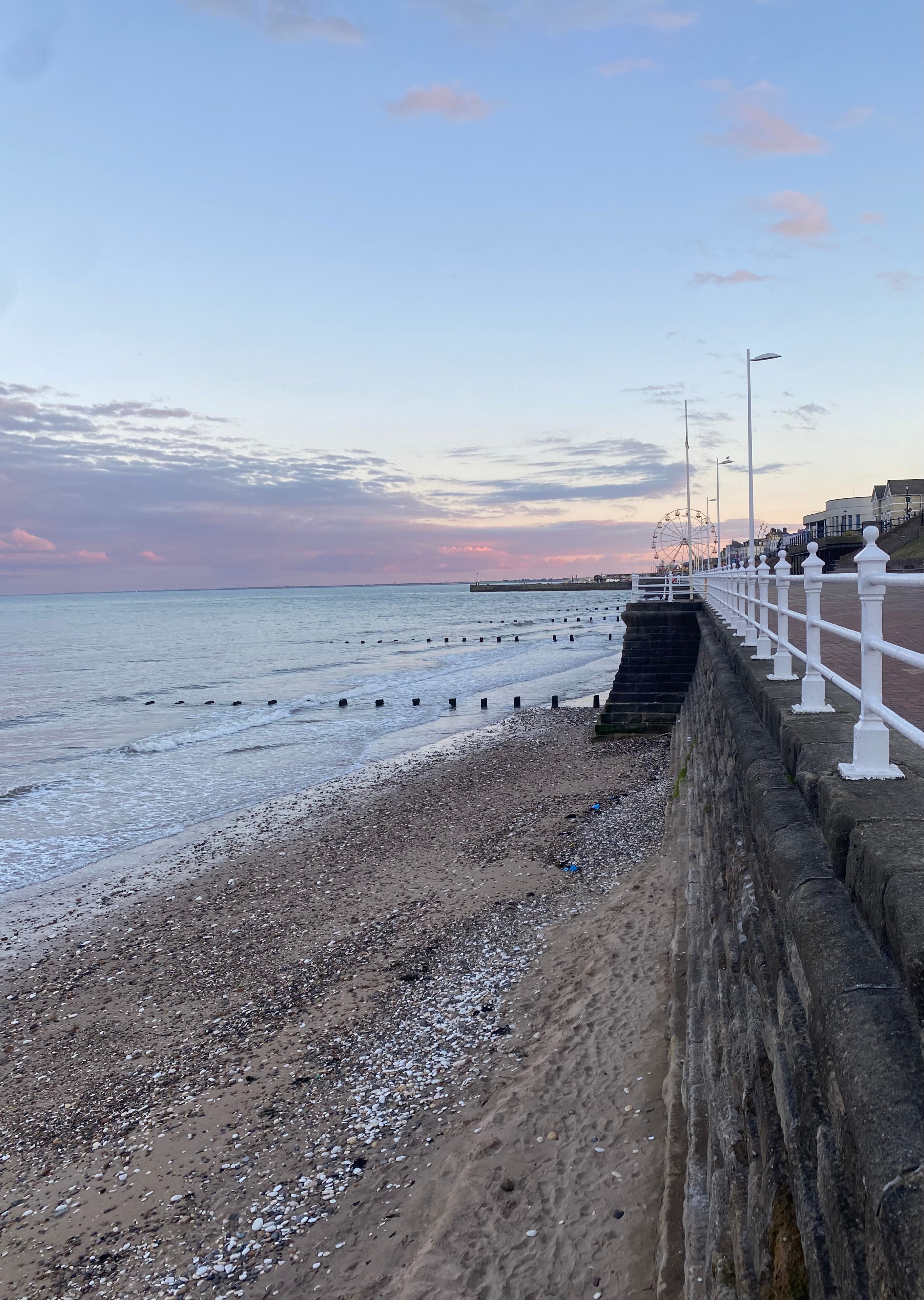 A scenic coastal view of Bridlington pebble beach and seaside promenade at sunset, with calm blue water, a pale sky with pink clouds, a stone wall, white railing posts, and a pier extending into the distance.