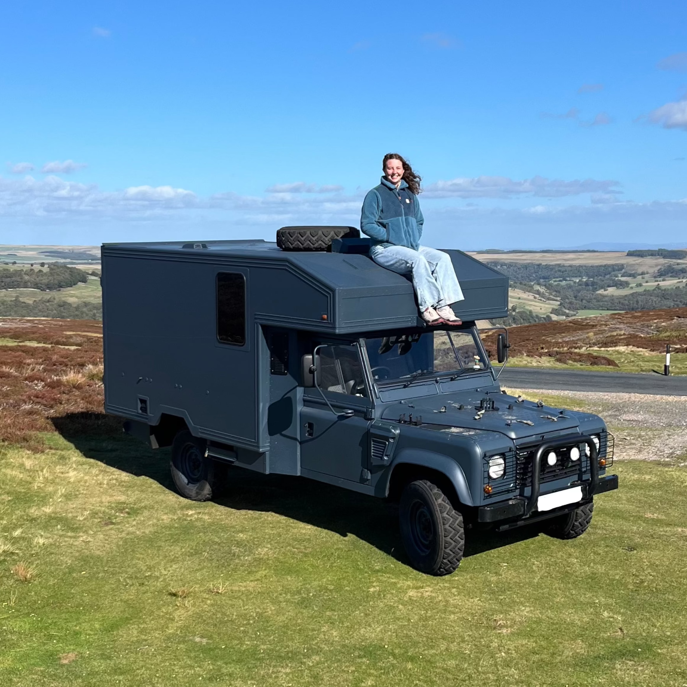 A woman sits on the roof of a navy blue Land Rover Defender 130 ex military ambulance conversion camper van parked on a grassy hillside with a wide-open landscape and blue sky in the background.
