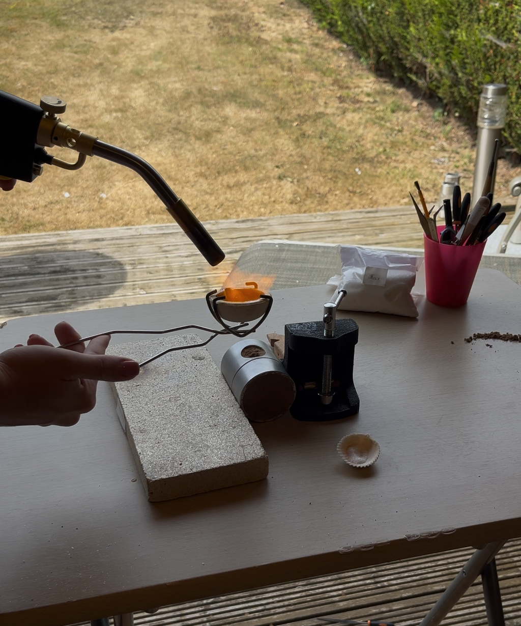 A hand holds a blow torch heating silver in a crucible about to pur into a sand cast mould on a workbench, with a heat block, tools in a pink cup, and an outdoor patio background visible.
