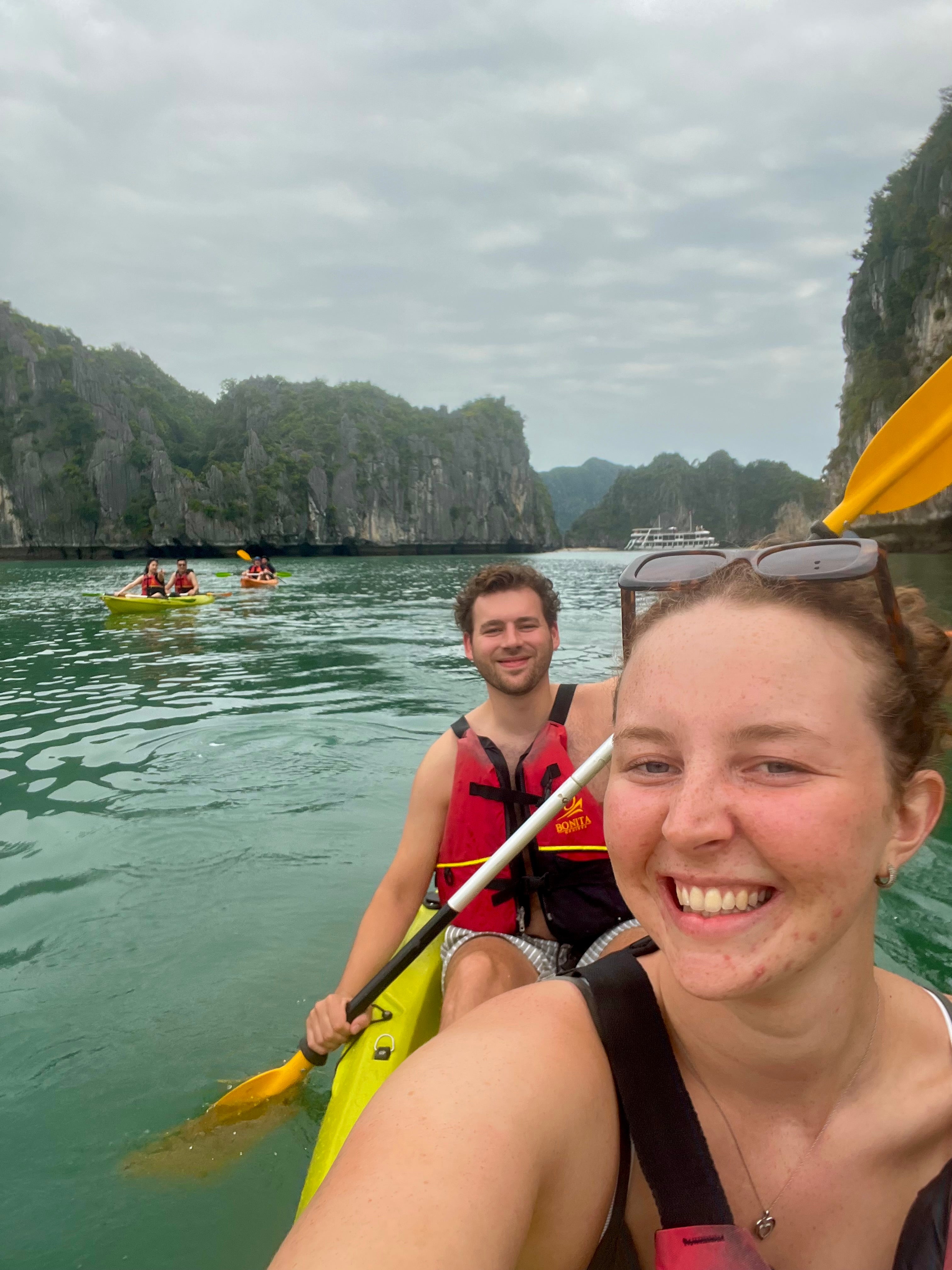 A woman in the foreground takes a selfie while wearing sunglasses on her head and holding a paddle, with a smiling man in a red life jacket behind her in a yellow kayak. They are on a body of water with tall green karst cliffs and other kayakers in the background under an overcast sky.