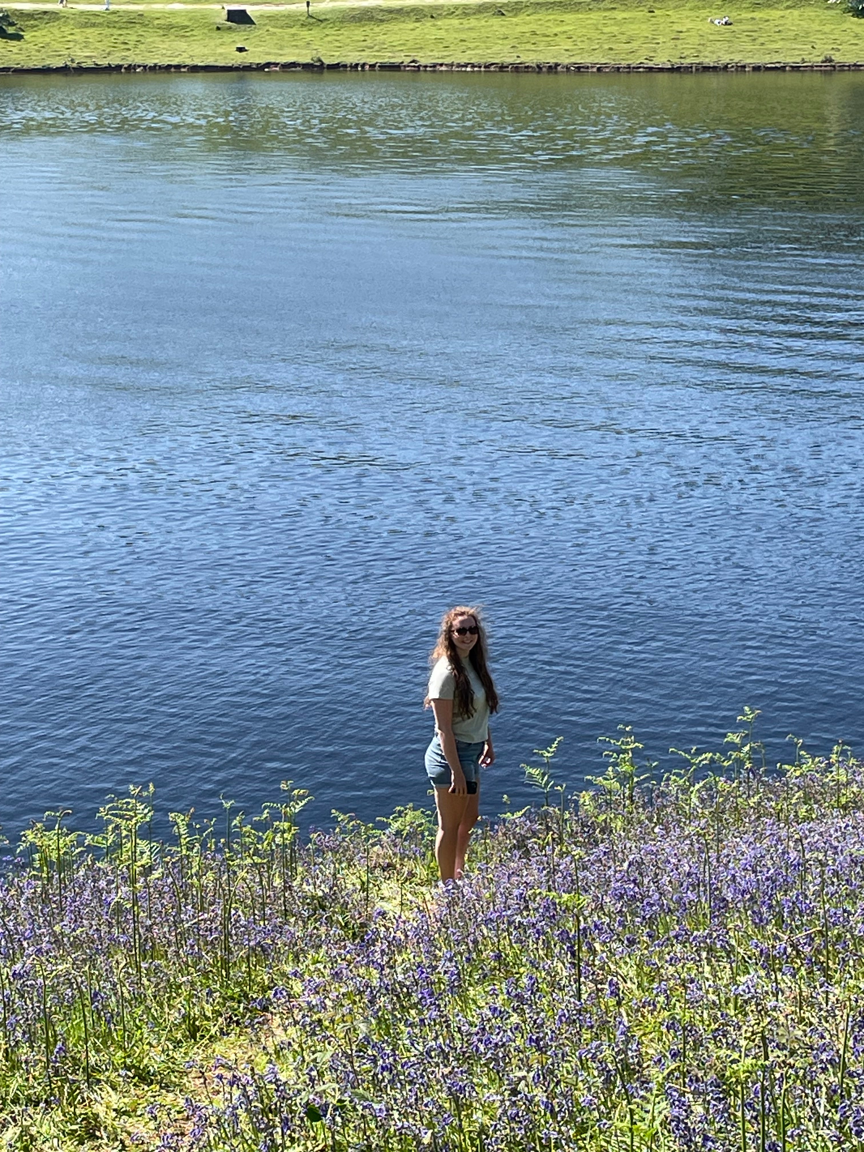 A woman stands on a grassy bank covered in purple wildflowers, looking out over a large blue lake or river under bright daylight.