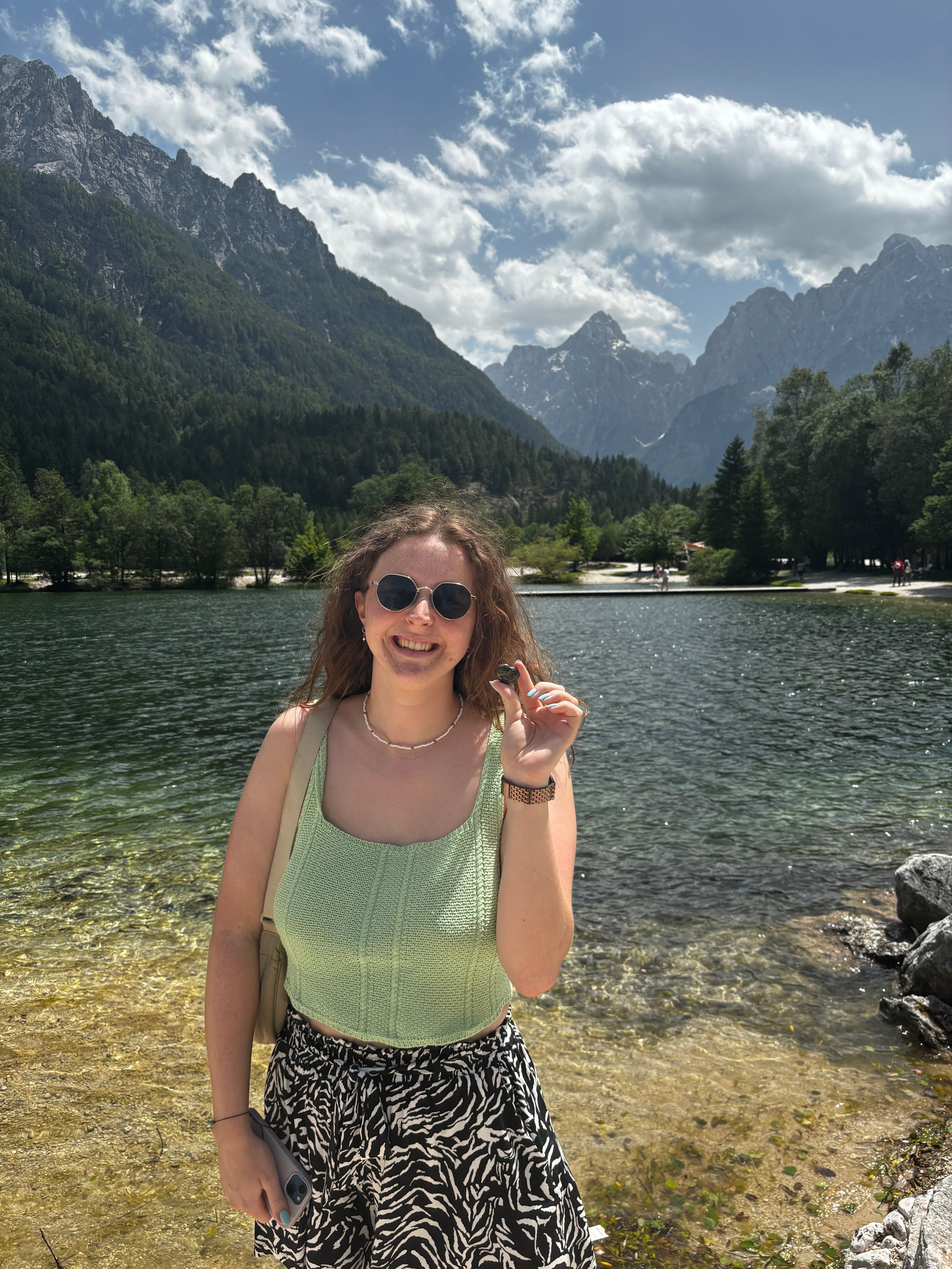 A woman posing outdoors in front of a mountain lake in Slovenia, wearing a light green knit tank top and a black-and-white patterned skirt.