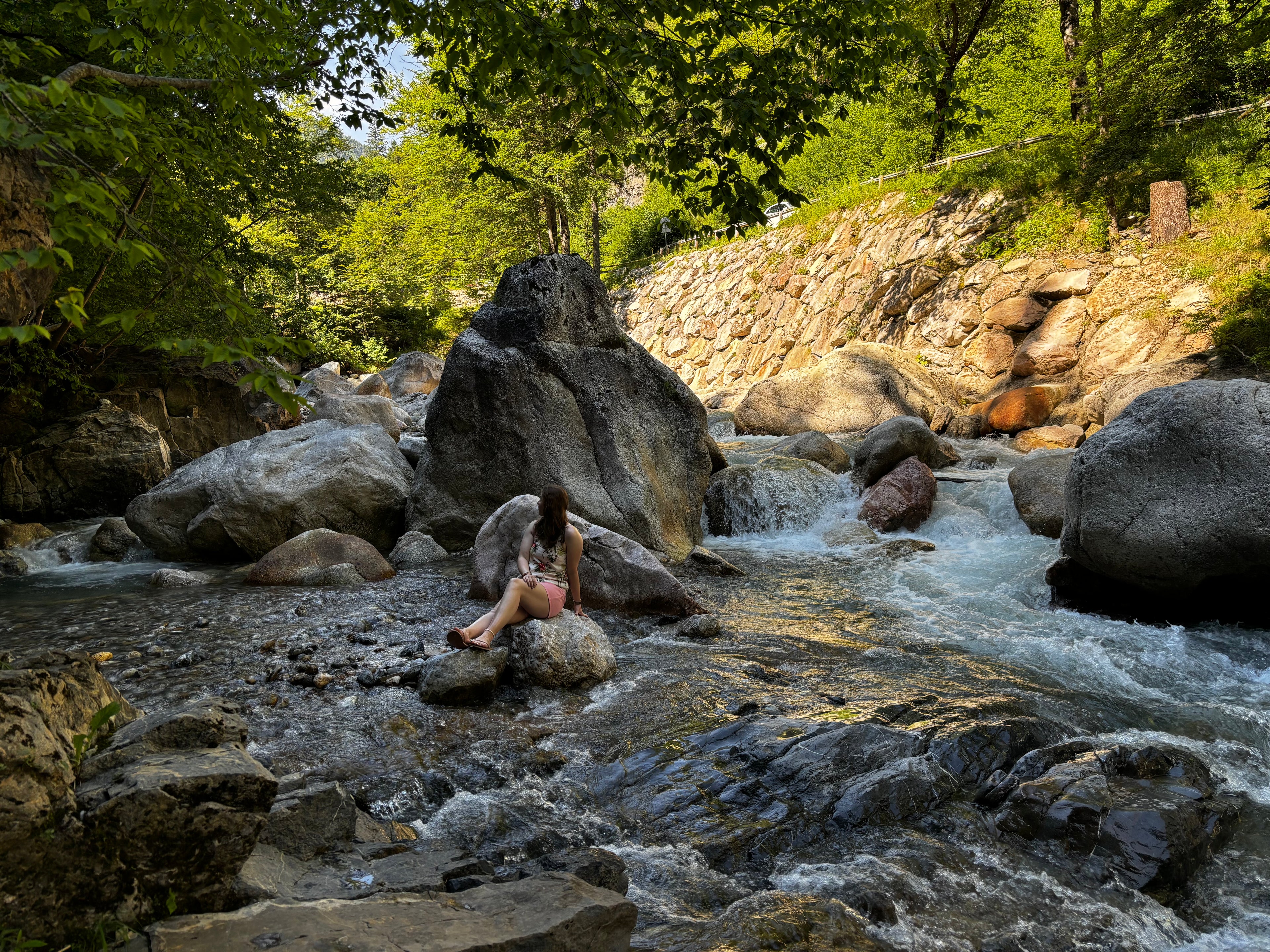 A woman sits on a large rock in a rocky stream in Slovenia surrounded by lush green trees and a stone wall in the background.
