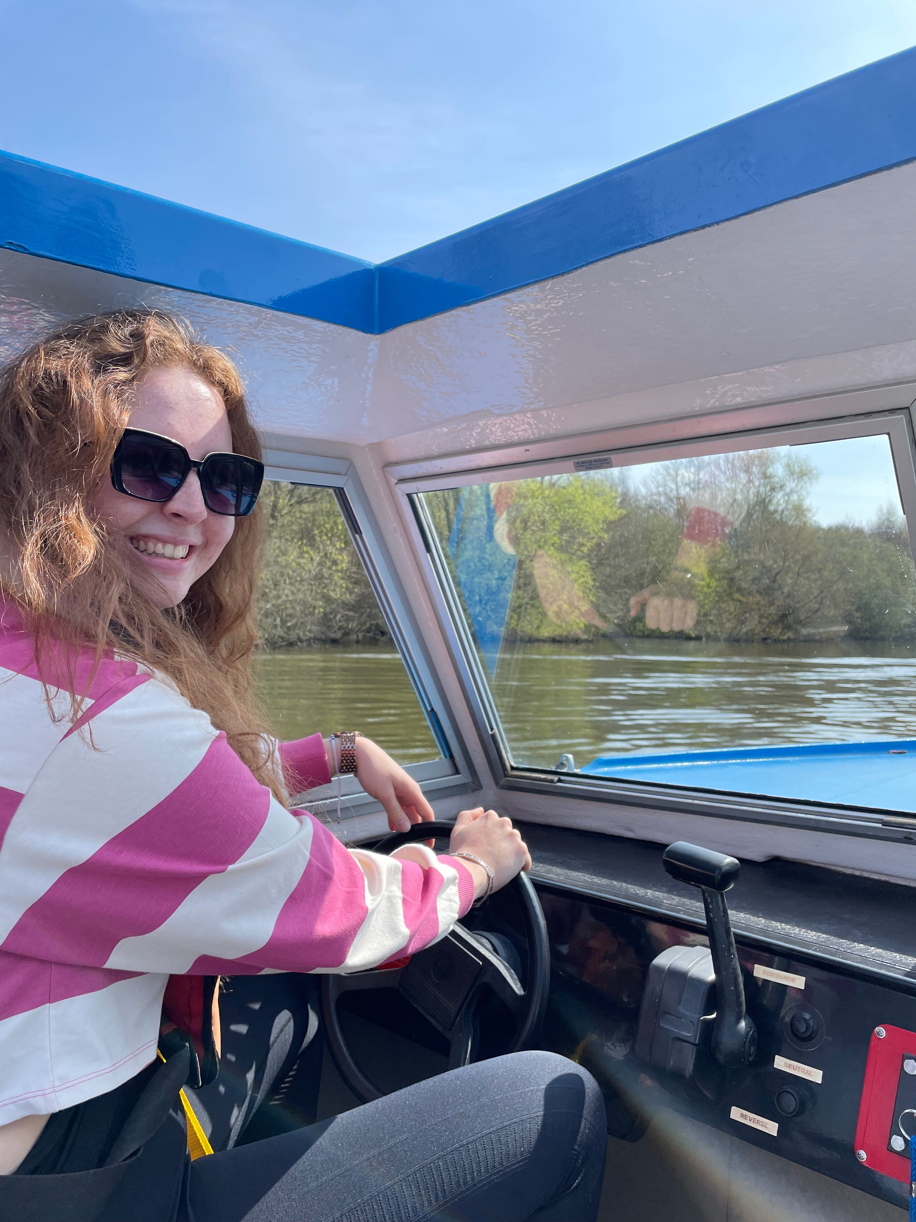 A woman wearing sunglasses and a pink and white striped shirt sits inside the cabin of a blue and white boat, holding the steering wheel with a river and trees visible outside.