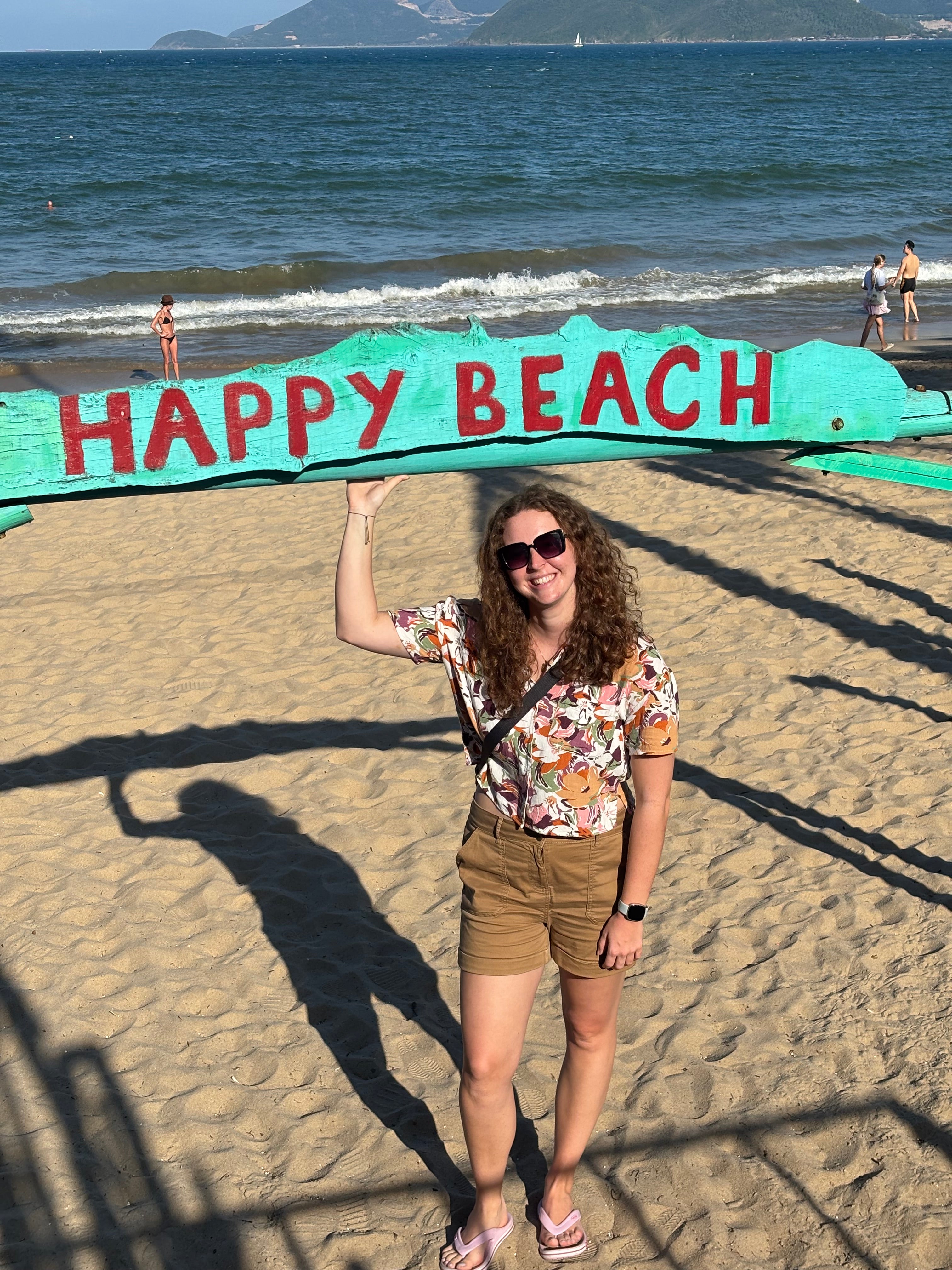 A woman posing on a sandy beach next to a large painted sign that reads 'HAPPY BEACH' in red letters on a teal background.
