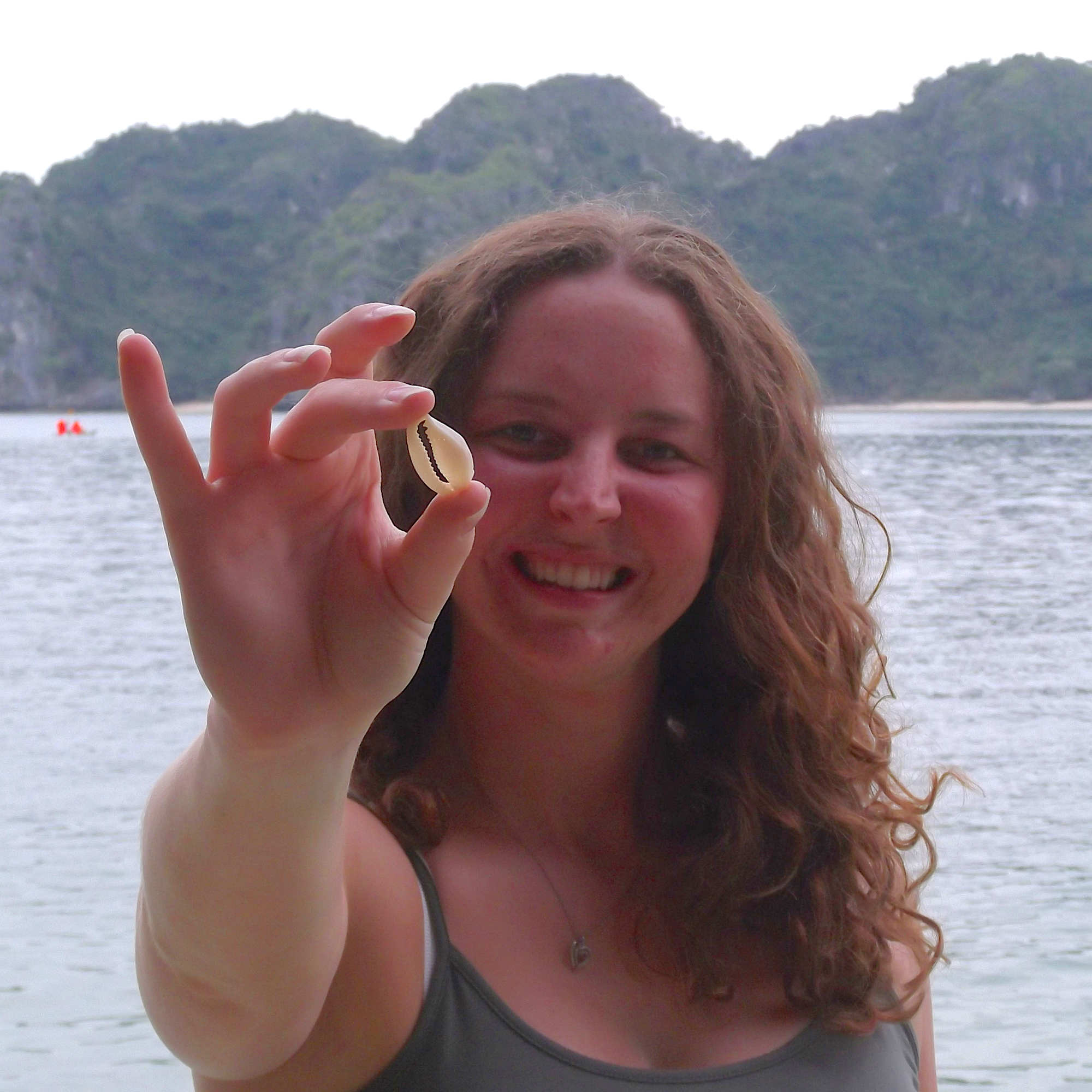A smiling woman with long wavy brown hair holds up a cowrie shell in front of her face, with the sea and green rocky cliffs in the background.