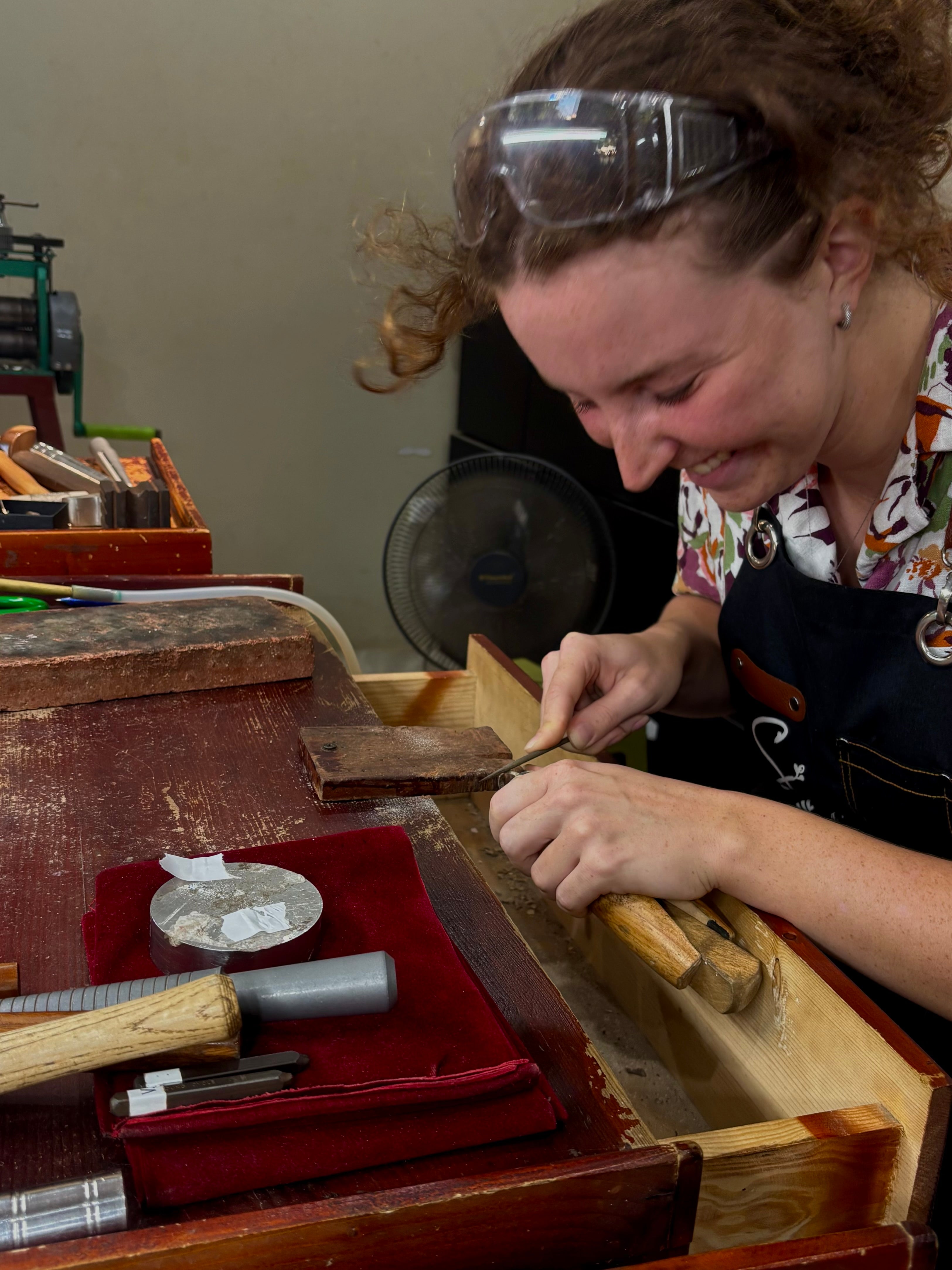 A woman wearing safety glasses and a dark apron smiles while working at a jewellery making bench, using a small hand tool to sand a ring held in a wooden vice. A red velvet cloth with tools is visible on the workbench in the foreground.