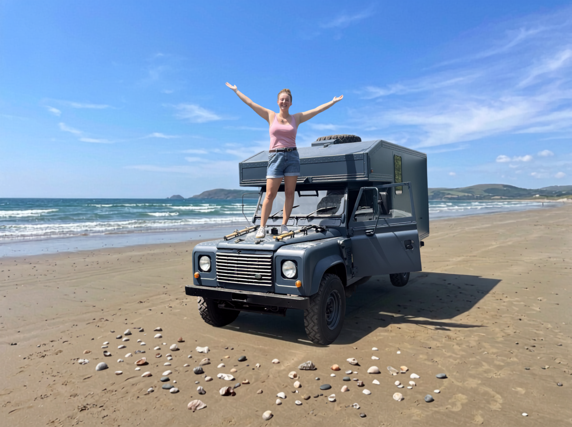 A woman stands with her arms raised on top of a Land Rover Defender 130 ex-military ambulance converted campervan parked on a sandy beach, with the ocean and blue sky in the background.