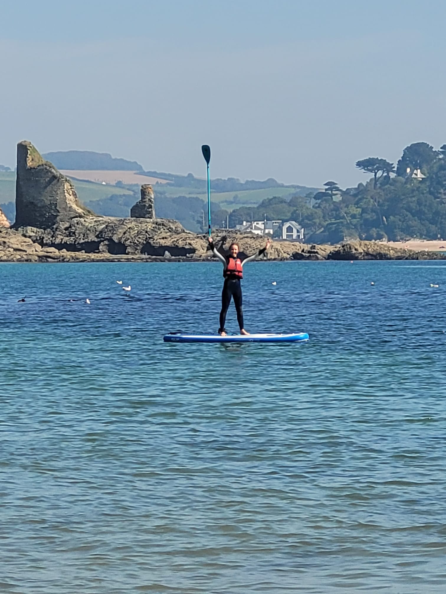 A person stands on a paddleboard in calm blue water with a paddle raised overhead, with rocky shoreline, trees, and a pale blue sky in the background.