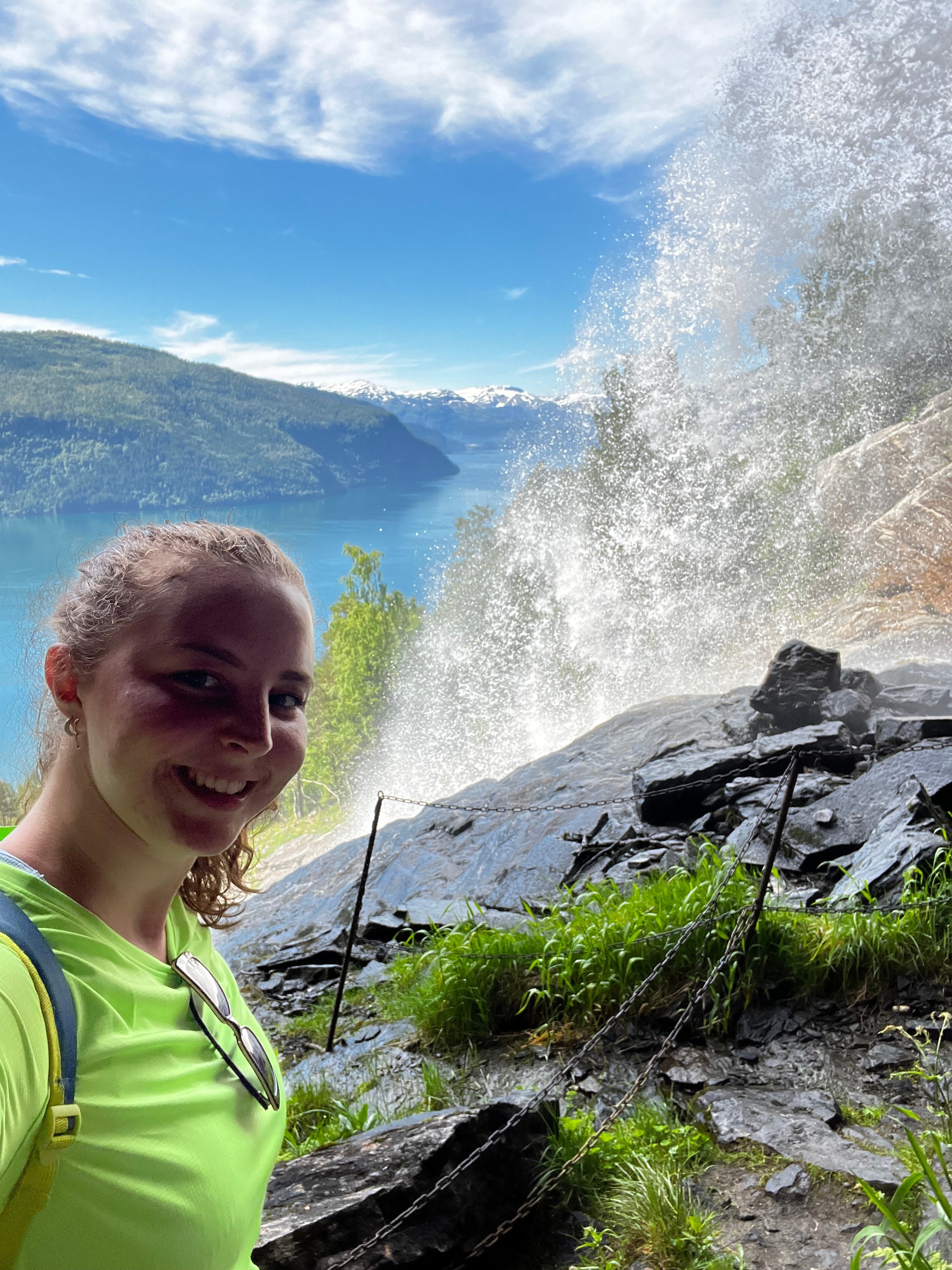 A woman in a bright lime green shirt takes a selfie in front of a dramatic waterfall cascading down a rocky cliffside next to a deep blue fjord and mountains under a blue sky in Norway.