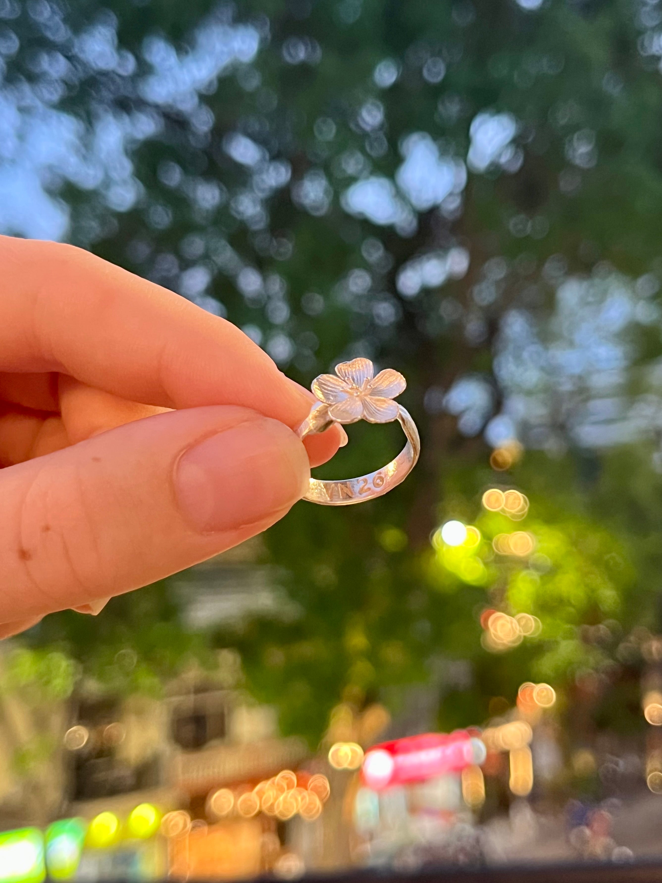 A hand holding a handmade silver ring with a small flower-shaped setting against a blurred outdoor background of green trees and warm bokeh lights.