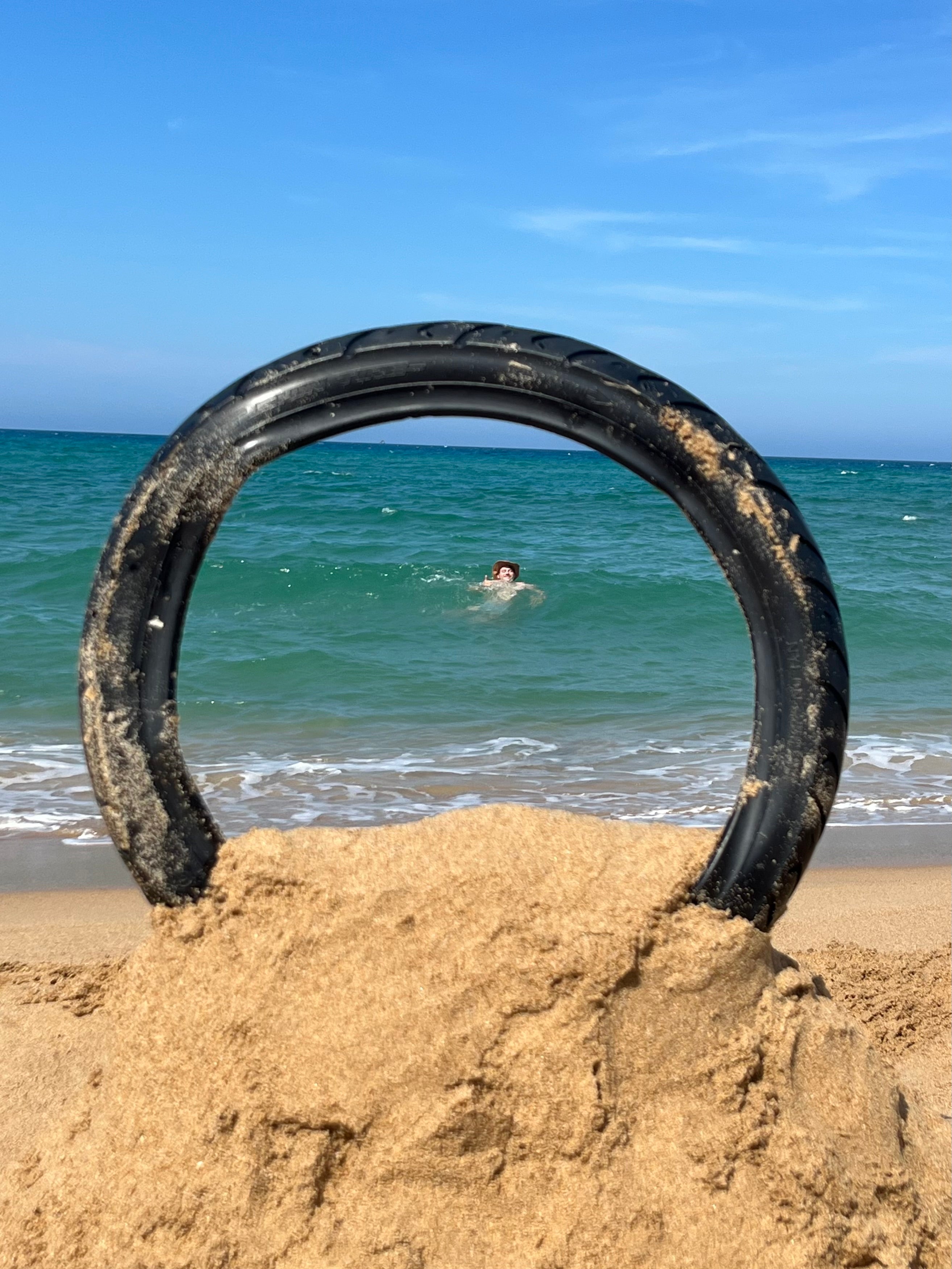 A black tire ring standing upright in a sandcastle on a beach, with the turquoise ocean and a swimmer visible in the background under a blue sky.